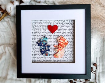 May include: Framed artwork featuring two origami figures, one blue and one orange, facing each other beneath a red heart. The background is a handwritten script on white paper, all within a black frame.