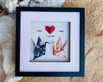 May include: Framed artwork featuring two origami cranes, one blue with a gold and white pattern, the other pink with a floral design. A red paper heart and the names "Rod" and "Steph" with the date "11.05.1974" are also included.