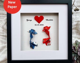 May include: A square black frame containing a white mat with two origami fish, one blue and one red, positioned below a red paper heart. The text George and Charlotte is printed on either side of the heart with the date 08.09.2024 printed below.
