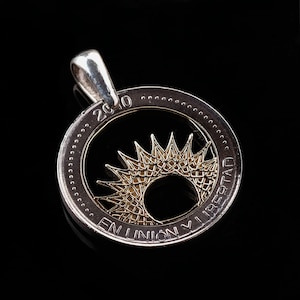 May include: A silver coin pendant with a gold sun design cut out of the center. The coin reads "2010 EN UNION Y LIBERTAD".