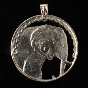 Puede incluir: Pendiente de plata con un diseño recortado de la cabeza de un elefante. El pendiente es detallado y tiene una superficie texturizada.