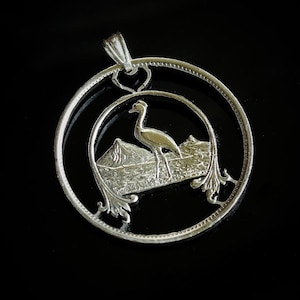 May include: Silver pendant featuring a crane with a mountain range in the background. The pendant is made from a coin with a cut-out design.