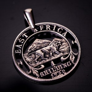 May include: Silver East Africa 1 Shilling coin from 1950 with a lion cut-out design. The coin has a loop at the top for hanging.