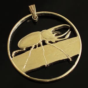 May include: A gold-toned pendant featuring a detailed cutout of a beetle with a large horn. The pendant is round and has a smooth, polished finish.