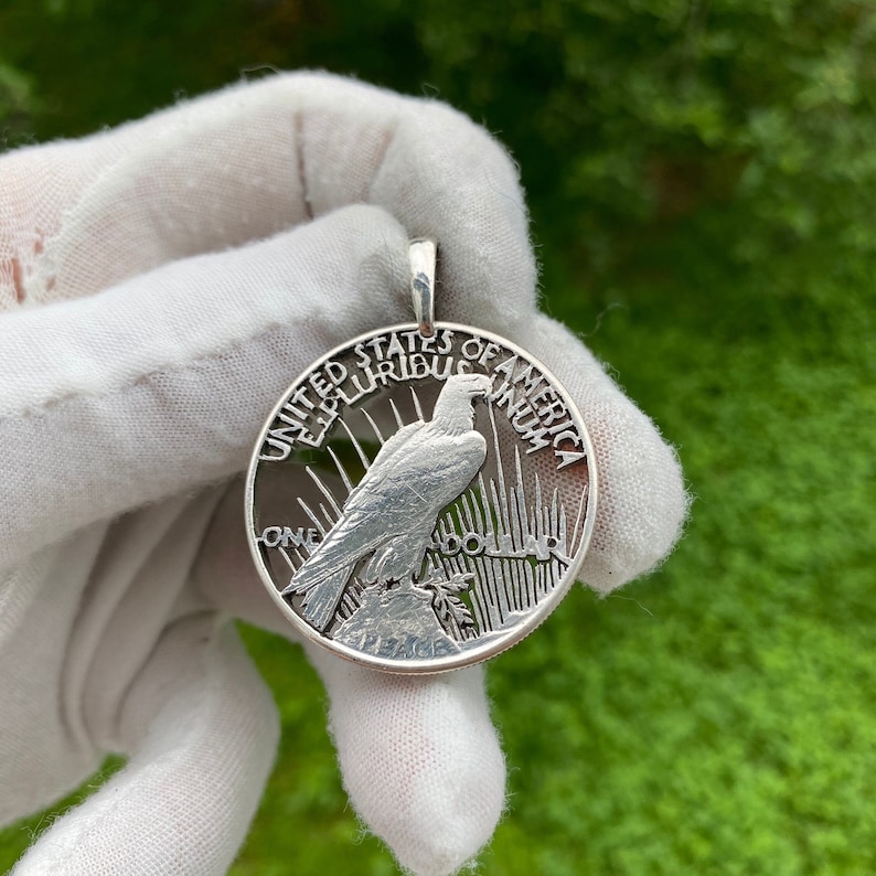 May include: A silver pendant featuring a detailed depiction of an eagle perched on a rock, with the words "UNITED STATES OF AMERICA" and "ONE DOLLAR" inscribed around the edge. The pendant has a loop for a chain.