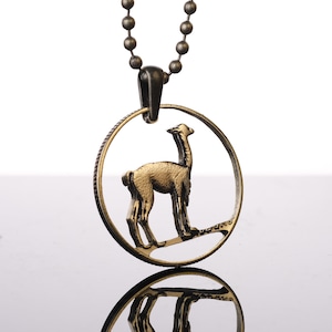 Puede incluir: Un collar de tono dorado con un colgante redondo que presenta un recorte de una llama. El colgante está unido a una cadena negra.