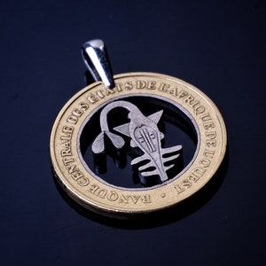 May include: A gold coin pendant with a cut-out design featuring a stylized image of a person's head. The coin is inscribed with the text "BANQUE CENTRALE DES ETATS DE L'AFRIQUE DE L'OUEST".