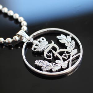 May include: A silver pendant necklace with a circular design. The pendant features a crown, the letter "R", and floral elements. The necklace has a ball chain and a silver clasp. The pendant is approximately 1.5 inches in diameter.