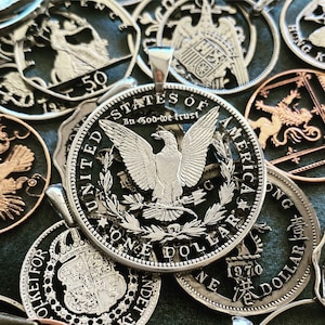 Pu&ograve; includere: Un ciondolo con una moneta da un dollaro americano argentata con un designo di aquila e la scritta "In God We Trust" e "One Dollar". La moneta &egrave; circondata da altre monete, tra cui una moneta da un dollaro di Hong Kong e una moneta britannica.