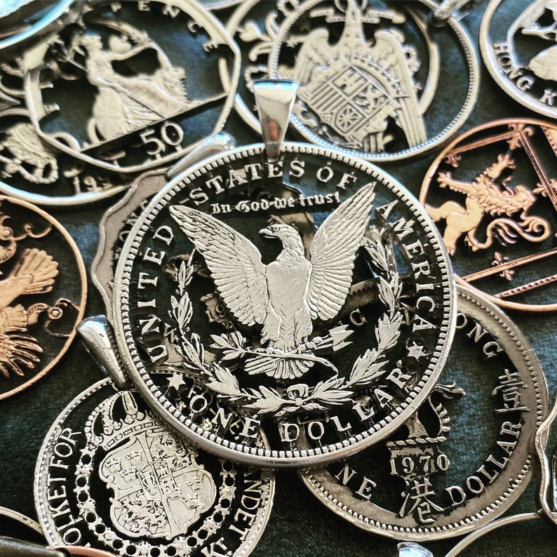 Pu&ograve; includere: Un ciondolo con una moneta da un dollaro americano argentata con un designo di aquila e la scritta "In God We Trust" e "One Dollar". La moneta &egrave; circondata da altre monete, tra cui una moneta da un dollaro di Hong Kong e una moneta britannica.