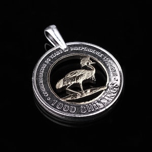 May include: A silver coin pendant with a cut-out design featuring a gold-colored bird. The coin reads "COMMEMORATING 50 YEARS OF INDEPENDENCE 1962-2012" and "1000 SHILLINGS".