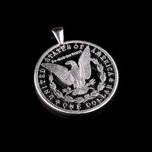 Pu&ograve; includere: Ciondolo a forma di moneta da un dollaro d'argento con un design a taglio di un'aquila con le ali spiegate. La moneta riporta la scritta "United States of America, In God We Trust, One Dollar".