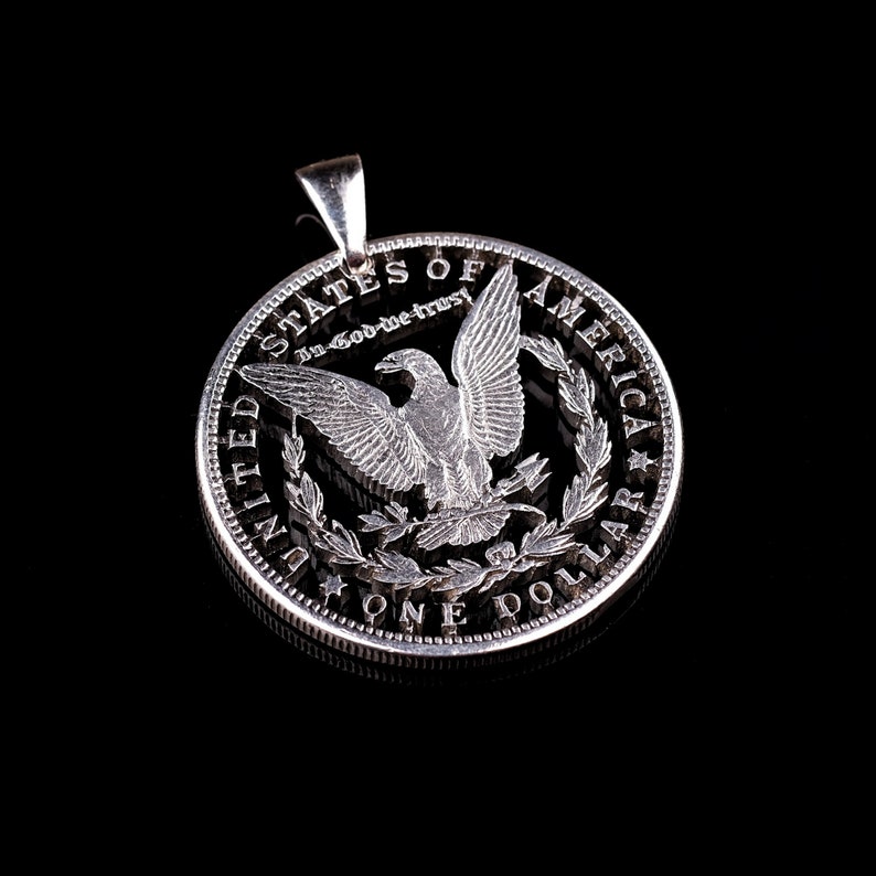 Pu&ograve; includere: Ciondolo a forma di moneta da un dollaro d'argento con un design a taglio di un'aquila con le ali spiegate. La moneta riporta la scritta "United States of America, In God We Trust, One Dollar".