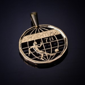 May include: A gold-colored pendant featuring a soccer player kicking a ball inside a globe with the word "ARGENTINA" and the number "78" on it.