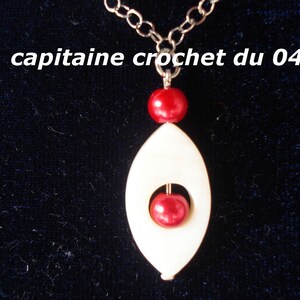 May include: A white pendant with a red bead in the center, hanging from a silver chain. The pendant is shaped like an oval with a hole in the center. The pendant is hanging against a dark blue background.