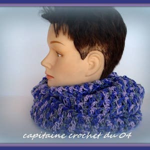 Peut inclure: Une écharpe en crochet infinie de couleur violette et bleue. L'écharpe est portée par un mannequin aux cheveux courts bruns.
