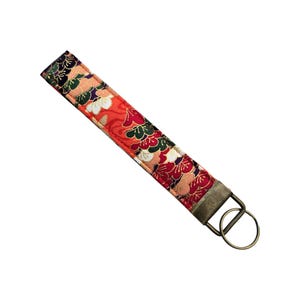 May include: A fabric wristlet key fob with a floral pattern in red, green, white, and gold. The key fob has a bronze-colored metal ring and hardware for attaching keys. The wristlet is approximately 6 inches long.
