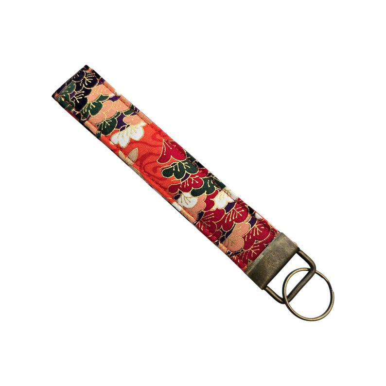 May include: A fabric wristlet key fob with a floral pattern in red, green, white, and gold. The key fob has a bronze-colored metal ring and hardware for attaching keys. The wristlet is approximately 6 inches long.