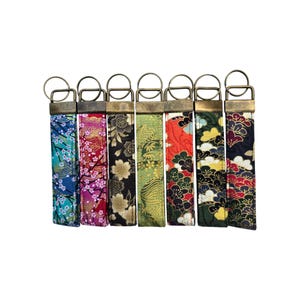 May include: Seven key fobs with brass-toned metal hardware and patterned fabric straps. The straps display floral and geometric designs in various colours, including blue, red, green, and black. Each fob has a metal ring for attaching keys.