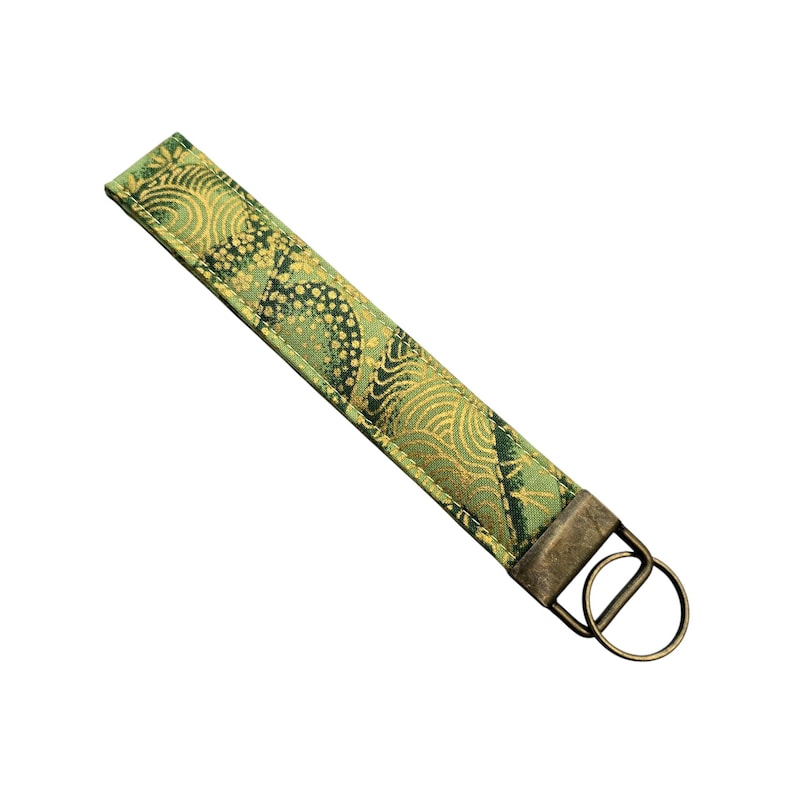 May include: A green and gold fabric wristlet key fob with a decorative pattern. The key fob has a rectangular shape with a metal ring and clasp for attaching keys. The fabric has a quilted appearance.