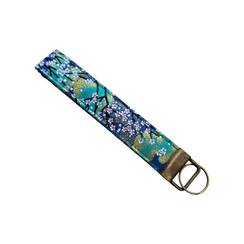 May include: A fabric wristlet key fob with a Japanese floral design. The key fob features a teal, navy blue, and gold pattern with white flowers and a bronze-colored metal ring.