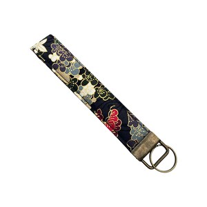 May include: A black fabric wristlet key fob with a floral pattern in white, red, gold, and blue. The key fob has a bronze-colored metal ring and hardware for attaching keys.