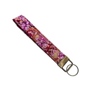 May include: A fabric wristlet key fob with a floral pattern in shades of red, pink, and gold. The key fob has a bronze-colored metal ring and clasp for attaching keys. The design features cherry blossom branches.