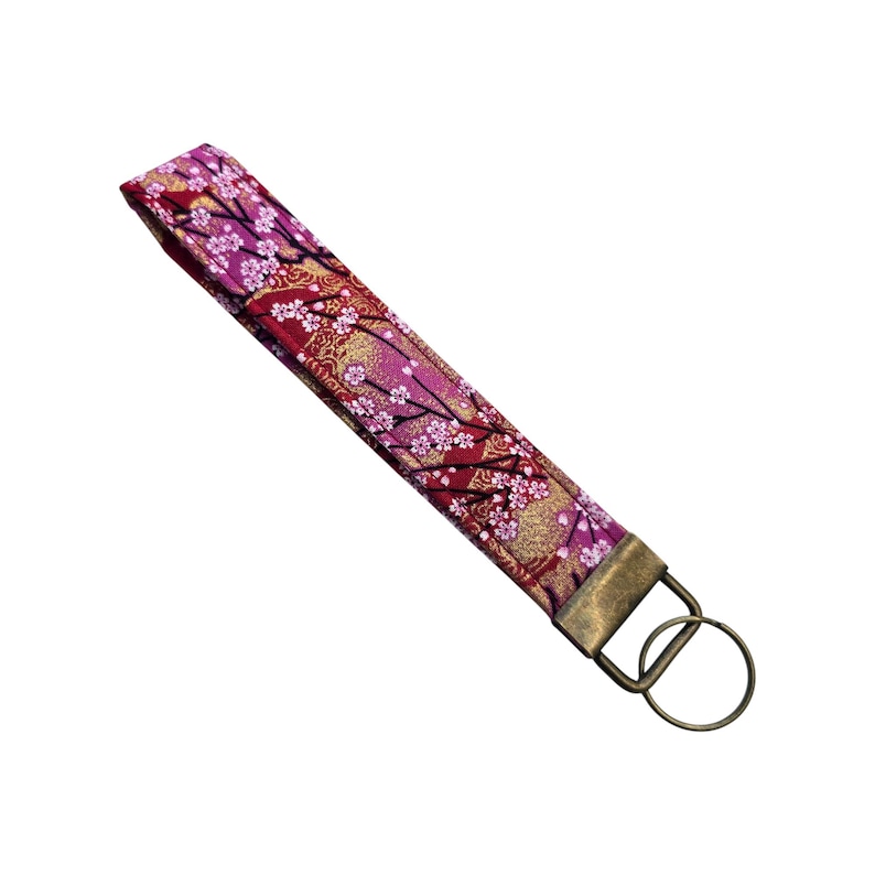 May include: A fabric wristlet key fob with a floral pattern in shades of red, pink, and gold. The key fob has a bronze-colored metal ring and clasp for attaching keys. The design features cherry blossom branches.