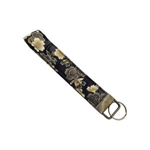 May include: A black and gold floral patterned wristlet key fob. The fabric features a repeating pattern of stylized flowers in shades of gold and gray against a black background. The fob has a metal ring and clasp for attaching keys.