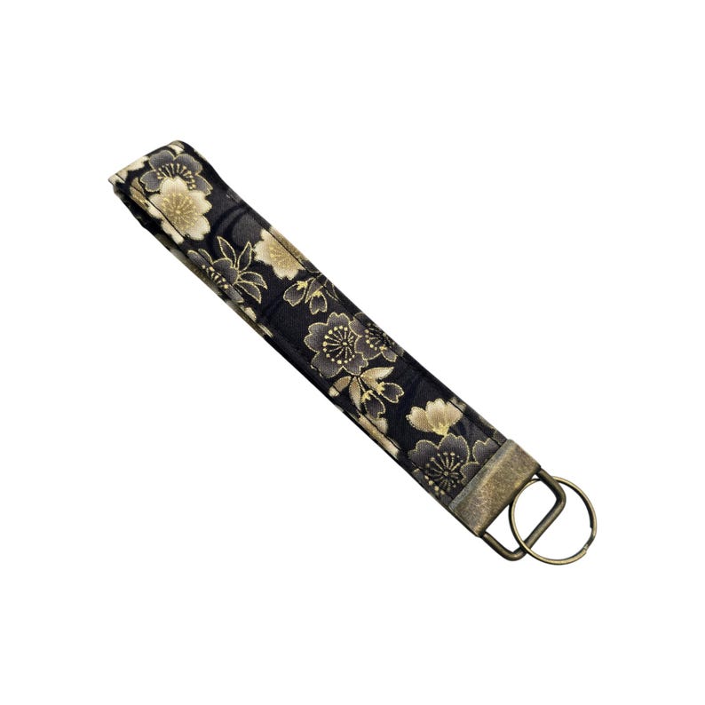 May include: A black and gold floral patterned wristlet key fob. The fabric features a repeating pattern of stylized flowers in shades of gold and gray against a black background. The fob has a metal ring and clasp for attaching keys.