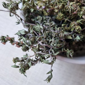 May include: Dried green herbs with small purple flowers in a white bowl.