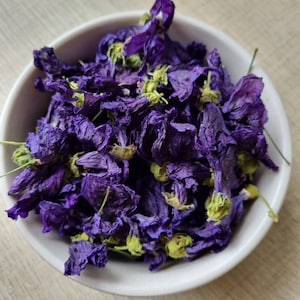 Può includere: Una ciotola di ceramica bianca piena di fiori viola secchi e boccioli verdi. I fiori hanno una consistenza rugosa e sono disposti a cerchio. La ciotola è appoggiata su una superficie di legno chiaro.