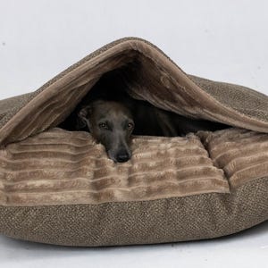 Puede incluir: Una cama para perros marrón con un exterior texturizado y un interior suave y afelpado. Un perro gris está acurrucado dentro de la cama, asomando por debajo de la capucha. La cama está diseñada para brindar comodidad y seguridad a los perros pequeños.