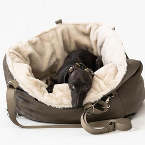 Op de afbeelding: Een donker olijfgroene draagtas voor huisdieren met een pluche, crèmekleurig interieur. Een kleine, zwarte hond ligt erin en kijkt recht in de camera. De tas heeft een schouderriem en metalen hardware.