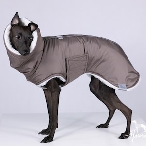 Op de afbeelding: Een bruine hondenjas met een witte fleece voering en een hoge kraag. De jas is ontworpen om een hond te passen en heeft een zak aan de zijkant. De hond is zwart en staat op een wit oppervlak.