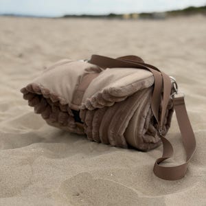 Puede incluir: Una manta beige enrollada con un borde de piel sintética texturizada y un exterior de tela lisa. La manta está asegurada con una correa beige a juego e incluye una correa de hombro ajustable de tejido resistente.