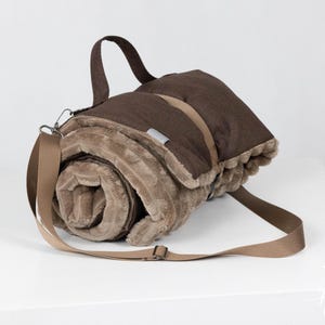 May include: A rolled-up brown and tan picnic blanket with a shoulder strap and carrying handle. The blanket is made of a soft, plush material and is secured with a matching strap. The blanket is designed for outdoor use.