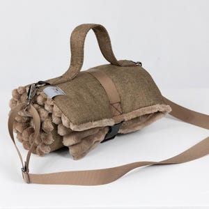 Può includere: Una coperta arrotolata per animali domestici, color beige, con manico e tracolla abbinati. La coperta ha un interno in peluche strutturato e un esterno liscio e tessuto. Una piccola etichetta è attaccata alla coperta.