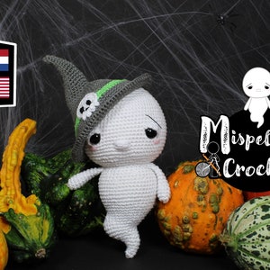 Op de afbeelding: Een handgemaakte witte gehaakte spookpop met een grijze heksenhoed met een schedel en groene accenten. Het spook wordt omringd door kalebassen en pompoenen, met een spinnenweb achtergrond. Een Halloween-thema.