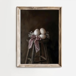 Op de afbeelding: Een ingelijste foto van drie witte eieren met een roze satijnen lint op een houten kruk. De eieren zijn in een groepje gerangschikt en omringd door kleine witte bloemen.