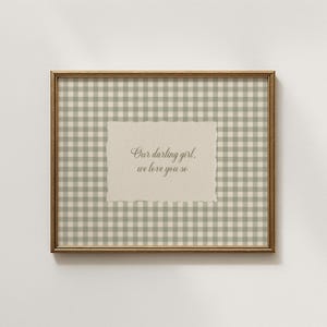 May include: A framed print with a gold-colored frame. The background features a green and white gingham pattern. A beige paper with the text "Our darling girl, we love you so" is centered on the print.