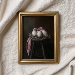 Op de afbeelding: Een ingelijst schilderij van drie witte eieren die in een krans van witte bloemen liggen, vastgebonden met een roze lint, op een houten kruk tegen een donkere achtergrond.