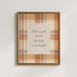 May include: A framed wall art print featuring a tan, brown, and blue plaid background with a central torn-edge paper overlay. The text reads: Fall is a gentle reminder that change can be beautiful. The frame is a thin, metallic gold color.