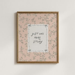 May include: Framed artwork with a gold-colored frame. The artwork features a pink floral patterned background. A white rectangle in the center has the handwritten text "JUST ONE MORE STORY". Small black bows are at each corner of the rectangle.