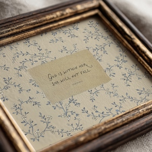 May include: Framed artwork featuring a floral pattern in blue and white on a beige background. A rectangular piece of paper with the text "God is within her, she will not fall. Psalm 46:5" is centered in the frame. The frame is a dark brown color.