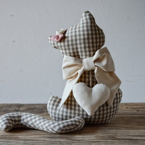 May include: A brown and white gingham cat-shaped doorstop with a white bow and a white heart. The doorstop is sitting on a wooden surface.