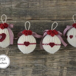 Può includere: Quattro palline di Natale in maglia bianca con cuori in feltro rosso e fiori in feltro rosso. Le palline sono decorate con un nastro a quadri rossi e bianchi.