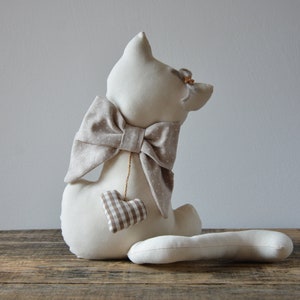 May include: A cream-colored stuffed cat with a large bow and a small heart-shaped charm. The bow is beige with white polka dots. The heart is brown and white checkered. The cat is sitting on a wooden surface.