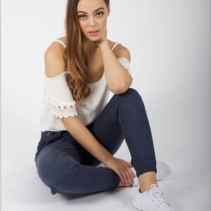 May include: A woman wearing a white off-the-shoulder top with lace trim and dark blue jeans. She is sitting on a white background with her legs crossed and her right hand resting on her neck.