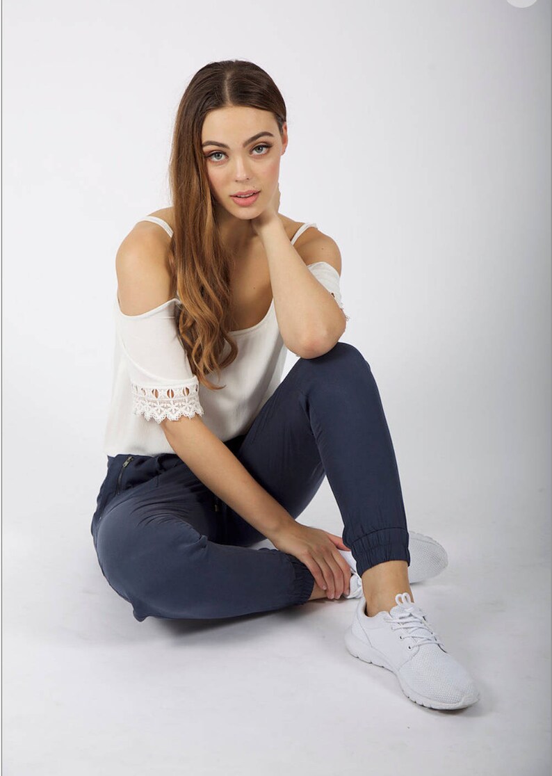 May include: A woman wearing a white off-the-shoulder top with lace trim and dark blue jeans. She is sitting on a white background with her legs crossed and her right hand resting on her neck.